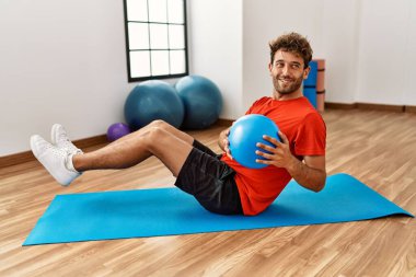 Genç İspanyol adam gülümsüyor spor merkezinde formda top kullanarak karın kaslarını çalıştırıyor.