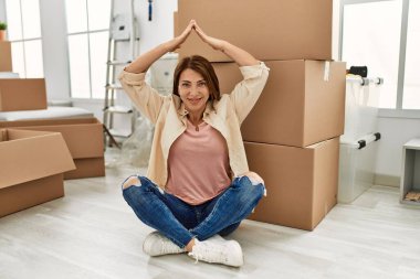 Orta yaşlı, beyaz bir kadın yeni evinde yerde elleri ile ev sembolü yapıyor..