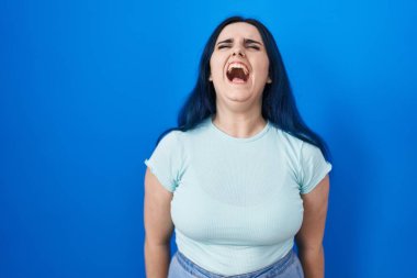 Young modern girl with blue hair standing over blue background angry and mad screaming frustrated and furious, shouting with anger. rage and aggressive concept. 