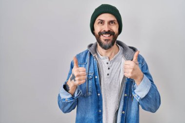 Yün şapka takan, elinde olumlu jest yapan, gülümseyen ve mutlu bir İspanyol dövmesi olan genç bir adam. Neşeli ifade ve kazanan jest. 