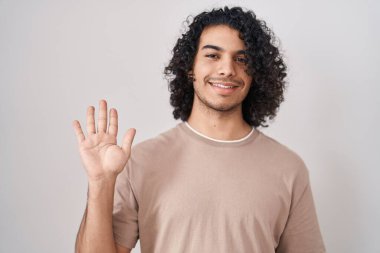 Beyaz arka planda duran kıvırcık saçlı İspanyol adam beş numarayı gösterip işaret ederken kendinden emin ve mutlu gülümsüyor.. 