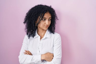 Hispanic woman with curly hair standing over pink background looking to the side with arms crossed convinced and confident 