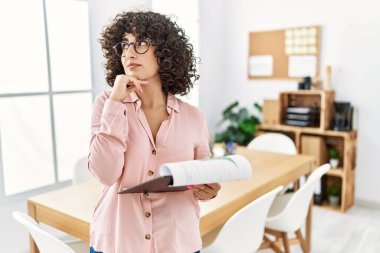 Ofiste iş elbisesi giyen genç bir ortadoğulu kadın elleri çenesinde soru sorarken, düşünceli bir ifade takınıyor. Düşünceli bir yüzle gülümsüyor. şüphe kavramı. 