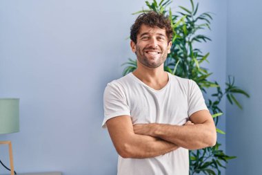 Genç adam kendinden emin gülümsüyor. Kollarını kavuşturup evde duruyor.