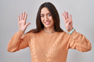 Beyaz arka planda duran genç esmer kadın dokuz numaralı parmaklarıyla işaret ederken kendinden emin ve mutlu bir şekilde gülümsüyor.. 