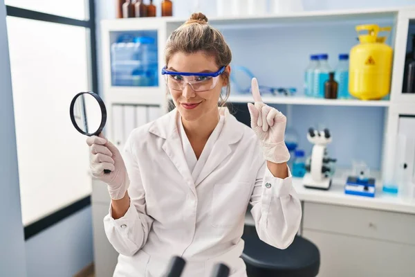 Young woman working at scientist laboratory holding magnifying glass smiling with an idea or question pointing finger with happy face, number one