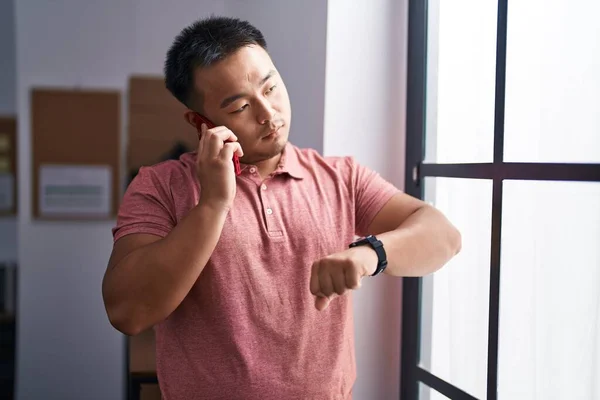 Young chinese man business worker talking on smartphone looking watch at office