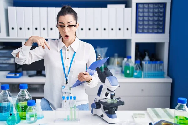 Young brunette woman working at scientist laboratory pointing down with fingers showing advertisement, surprised face and open mouth