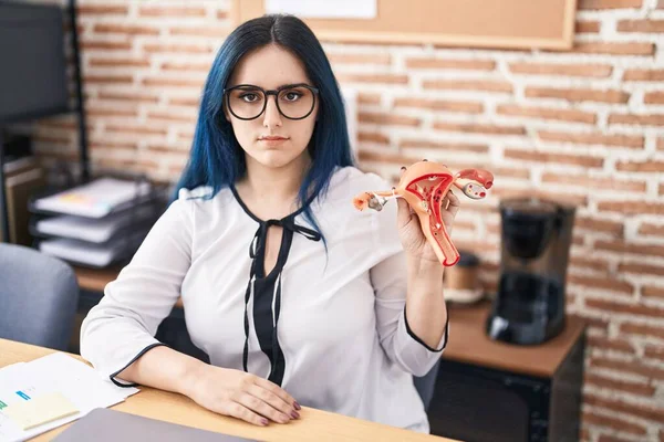 Young girl with blue hair holding model of female genital organ at the office complaining for menstruation pain thinking attitude and sober expression looking self confident