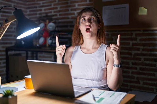 Brunette woman working at the office at night amazed and surprised looking up and pointing with fingers and raised arms.