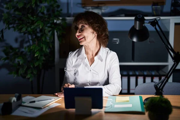 Middle age woman working at the office at night looking away to side with smile on face, natural expression. laughing confident.