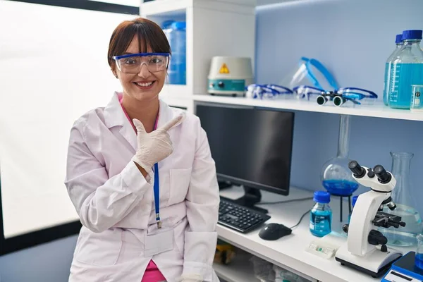 Young brunette woman working at scientist laboratory cheerful with a smile of face pointing with hand and finger up to the side with happy and natural expression on face