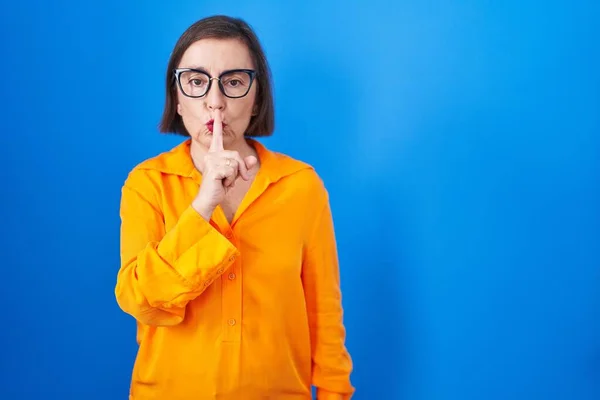 青い背景の上に眼鏡をかけた中年のヒスパニック系女性が唇に指で静かにするように求めている 沈黙と秘密の概念 — ストック写真