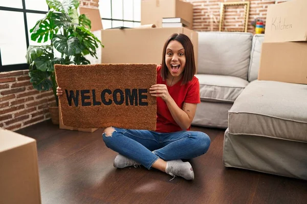 Young hispanic woman holding welcome doormat at new home celebrating crazy and amazed for success with open eyes screaming excited.