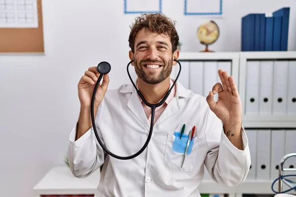 Young hispanic doctor man wearing doctor uniform holding stethoscope at clinic doing ok sign with fingers, smiling friendly gesturing excellent symbol