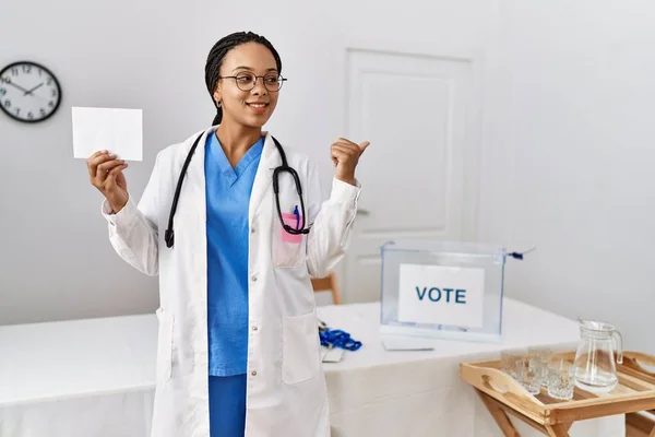 Young african american doctor woman voting holding envelope pointing thumb up to the side smiling happy with open mouth