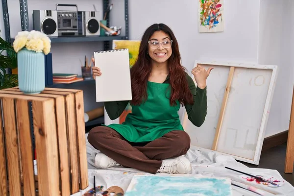 Hispanic Young Woman Holding Notebook Art Studio Pointing Thumb Side — Foto de Stock