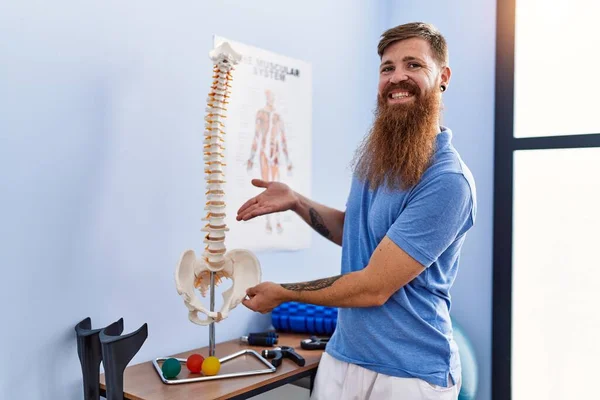 Young redhead man wearing physiotherapist uniform pointing to anatomical model of vertebral column at physiotherapy clinic