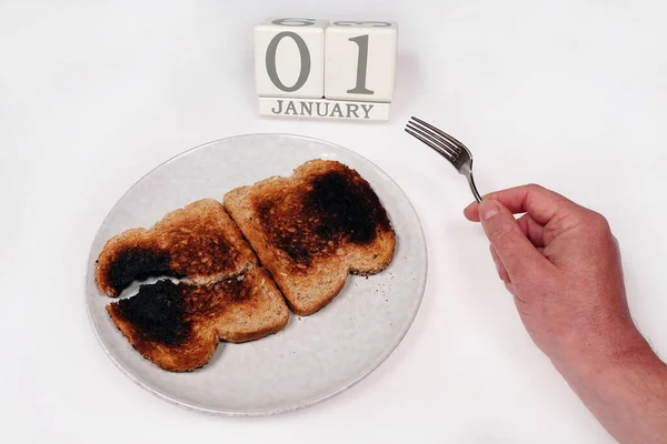 New Year starts with Poverty. Black burned toast on a plate with hand, fork and date of January 1st isolated on white background.