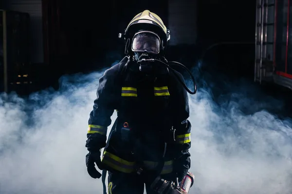 Portrait of a female firefighter wearing a helmet and all safety equipment a while holding a tomahawk and wearing an oxygen mask indoors surrounded by smoke