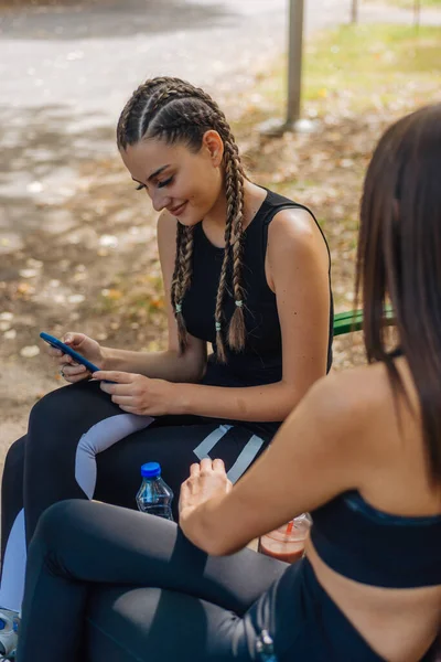 公園に座ってスポーツ活動から休憩している間に 魅力的な女性が笑顔で携帯電話をスワイプします — ストック写真