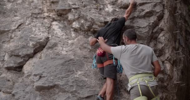 彼の友人が彼を助けている間ハンサムなシニア男性は登山者のための大きな岩を準備するために登山しています — ストック動画