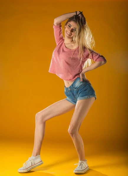 タンクトップのスポーティ美少女と喜びと笑顔で踊るデニムショートパンツ 黄色の背景に手を振ってヴィンテージヘアスタイルと見事な女性モデルの屋内肖像画 — ストック写真