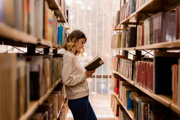 College Mädchen Liest Ein Buch Während Sie Zwischen Den Bücherregalen — Stockfoto