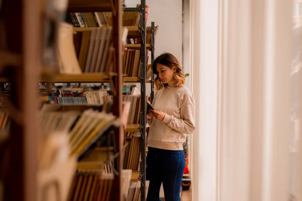 College Girl Reading Book While Standing Bookshelves — Stockfoto