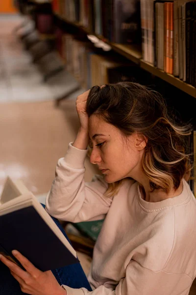 女孩在图书馆的书架上看书 一边坐着一边挽着头发 — 图库照片
