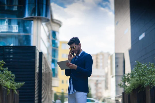 Urban businessman is smiling while talking on the phone and looking at his ideas and plans on his tablet