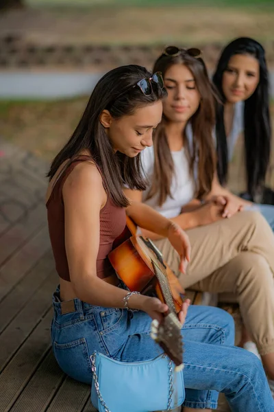 beautiful girl friends are listening to their friend play the guitar while sitting on the bench