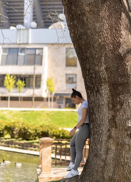 春の晴れた日に公園で木にもたれかかる暗い髪とカジュアルな服装の若い女性 — ストック写真
