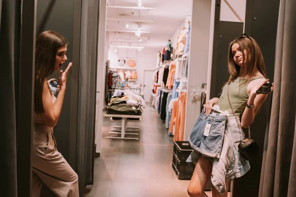 Two pretty girls standing at the changing room in the shop trying clothes and showing them to each other