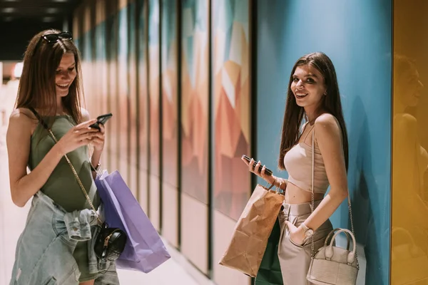 Two beautiful best friends looking at their phones while standing in the hallway at the mall