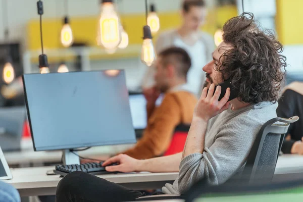 Bearded employee guy with afro haircut talking on his phone while being surrounded by his multiethnic coworkers. Businessman working in a casual startup modern environment.