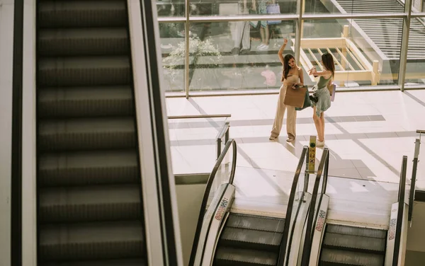 两个漂亮的姑娘站在市区购物中心的自动扶梯旁 一边拿着电话一边购物 — 图库照片