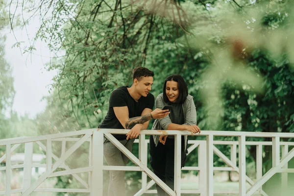 公園の白い橋にもたれながら かわいいカップルの電話を見て閉じる — ストック写真