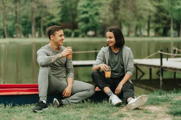 在湖边的公园里 一对美丽的夫妇坐在一只红色的皮划艇上 手里拿着果汁 — 图库照片