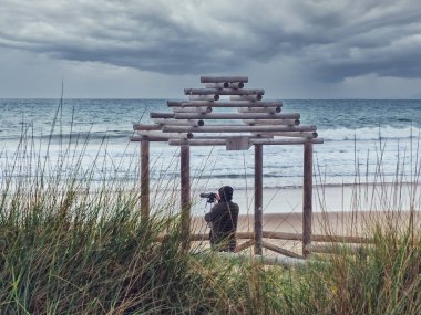 Kasvetli havada yalnız bir fotoğrafçı ahşap bir gölgeliğin altında denizi fotoğraflıyor..
