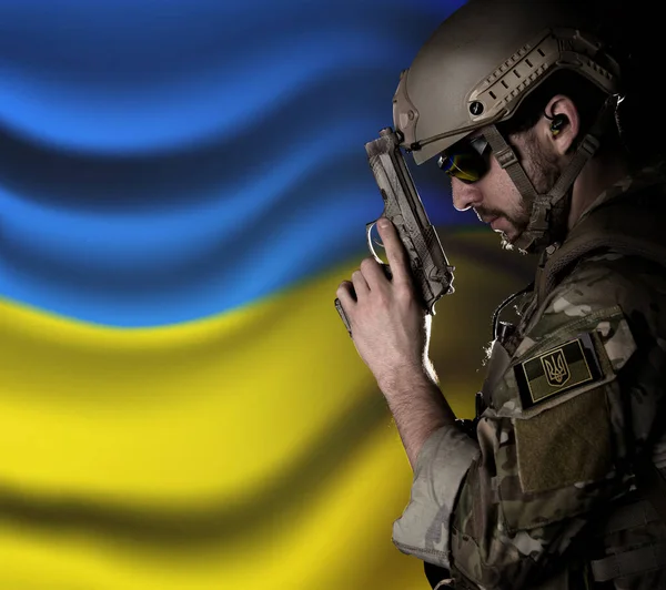 Ukrayna askeri kamuflaj üniforması içinde, Ukrayna arması ile Ukrayna bayrağının arka planında, düşünceli bir pozda, silahlı. Askeri çatışma
