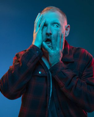 Mavi stüdyo arka planında kırmızı cyan neonlu, çok renkli duygusal bir adamın portresi. Çok büyük bir korkuyla, elleriyle yüzünü tuttu ve parmaklarıyla yan tarafına baktı. Broşür Posteri ReklamcıIığı