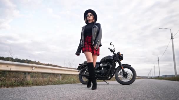 Sexy motorcyclist woman in mini skirt, leather jacket, hat walking to camera on retro-styled motorcycle background. Attractive female driver in jackboots on highway. Trip, speed, freedom concept. — Stock Video