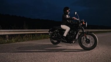 Motosikletli kadın retro-stil motosikletin üzerinde oturuyor. Gece yolda kask takan genç bir kadın sürücü. Trip, özgürlük, klasik motosiklet konsepti.
