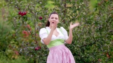 Dirndl 'deki komik kadın - geleneksel festival elbisesi komik dans ediyor ve olgun kırmızı elmalı sonbahar bahçesi arka planında şarkı söylüyor. Dans, maskaralık, pozitif ruh hali ve eski tarz konsept.