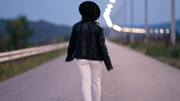 Young woman walking from camera on empty night street with lanterns. Girl in white pants and leather jacket. Hipster in hat. Stylish lady goes into distance — Stock Video