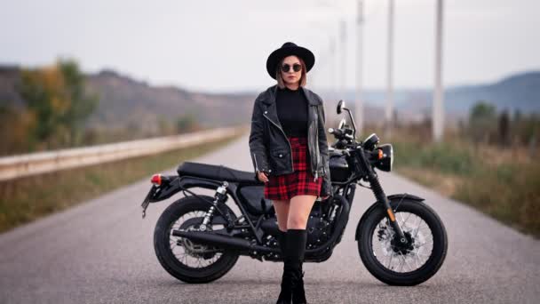 Mujer sexy en falda de cuadros rojos, chaqueta de cuero y sombrero caminando a la cámara en el fondo de la motocicleta de estilo vintage. Atractiva motociclista femenina con botas en la carretera. Viaje, velocidad, concepto de libertad. — Vídeos de Stock