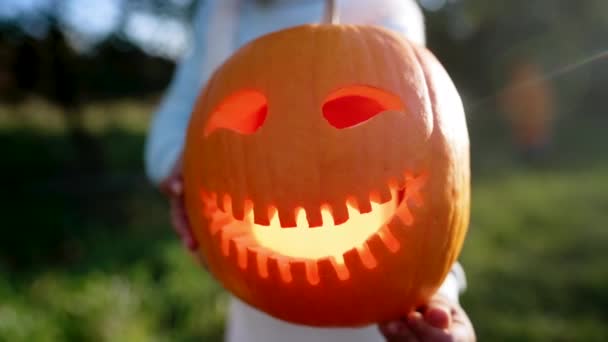 Dziecko trzyma rzeźbioną uśmiechniętą dynię na słonecznym tle ogrodu. Halloween, cukierek albo psikus. Symbol Wszystkich Świętych Wigilii — Wideo stockowe