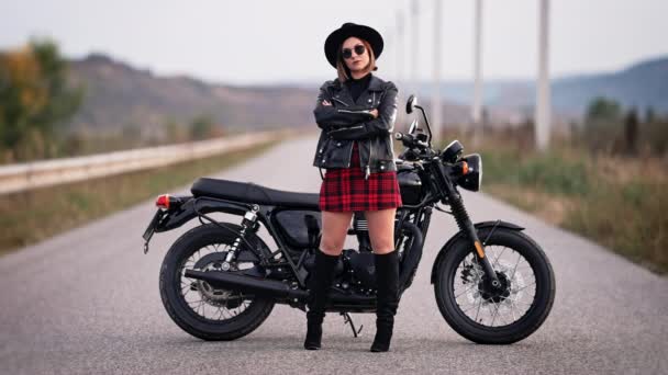 Portret motocyklistki w mini spódnicy, skórzanej kurtce i kapeluszu stojącej w pobliżu zabytkowego motocykla. Kierowca w butach i okularach słonecznych na jezdni. Podróż, szybkość, styl, koncepcja feminizmu. — Wideo stockowe