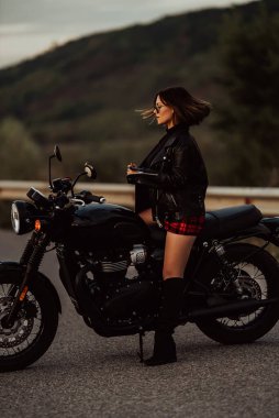 Kendine güvenen motosikletli kadın portresi mini etekli, deri ceketli retro-stil motosikletli. Otobanda çizmeli çekici bir sürücü. Trip, hız, tarz, feminizm kavramı.